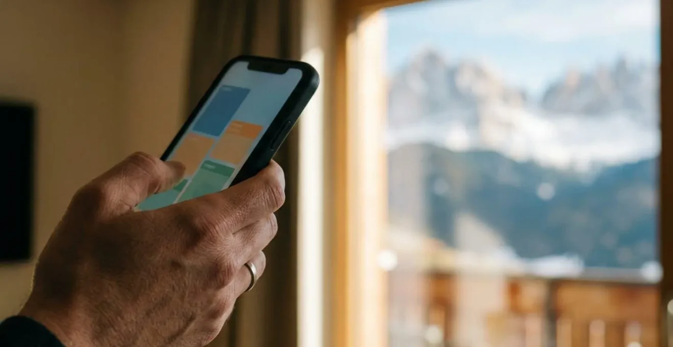 Mano che tiene smartphone mostrando calendario colorato con prezzi skipass variabili per date diverse, sfondo sfocato di montagne innevate