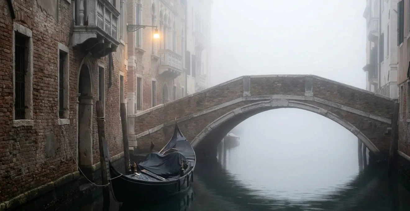 Canale veneziano nella nebbia invernale con un ponte e una gondola solitaria